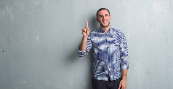 年轻的白种人男子在灰色的格朗格墙上展示和指向第一手指，同时微笑自信和快乐。