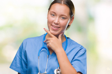 年轻的白种人医生女士穿着医疗制服，在孤立的背景下，自信地看着相机，微笑着交叉的手臂和举起的手在下巴上。 积极思考。