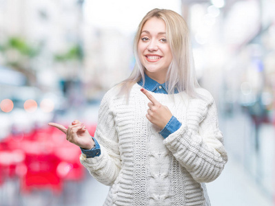 年轻的金发女人穿着冬天的毛衣，在孤立的背景上微笑着，看着镜头，双手和手指指向一边。