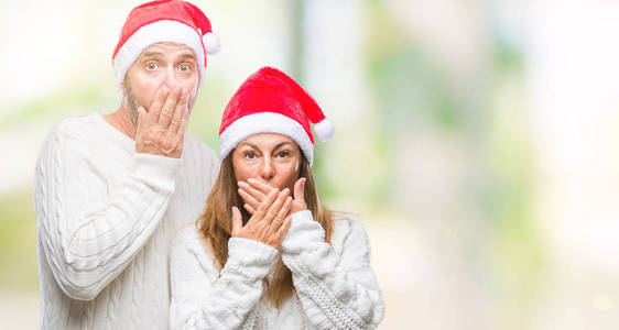 中年西班牙裔夫妇戴着圣诞帽，在孤立的背景下震惊地用手捂住嘴，因为他们弄错了。 秘密概念。