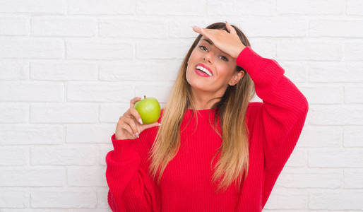 年轻的成年女子在白色砖墙上吃着新鲜的绿色苹果，用手压着头，羞愧和惊讶的脸，愤怒和沮丧。 因为错误而恐惧和不安。