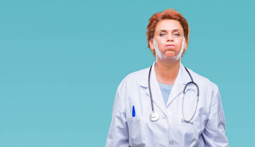 高级白种人医生女士穿着医疗制服，在孤立的背景下，鼓着脸颊，带着滑稽的脸。 嘴里充满了空气疯狂的表情。