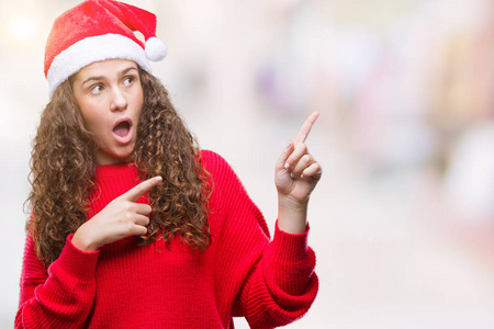 年轻的黑发女孩戴着圣诞帽在孤立的背景上微笑着，看着相机，两只手和手指指向一边。