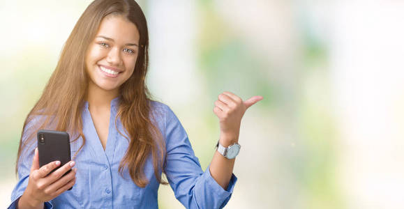 年轻漂亮的黑发商务女性使用智能手机，在孤立的背景下指着并用拇指向侧面展示，脸上洋溢着幸福的笑容