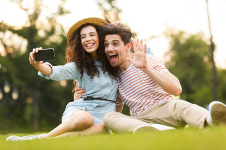 20多岁的漂亮情侣坐在公园的草地上，在智能手机上自拍