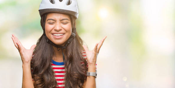 年轻的阿拉伯骑自行车的女人戴着安全帽，在孤立的背景下庆祝疯狂和疯狂的成功，举起手臂，闭上眼睛，尖叫兴奋。赢家概念