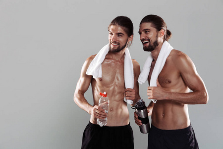 两个笑容可掬肌肉发达赤膊上阵的孪生兄弟运动员站在灰色背景下，手里拿着水瓶