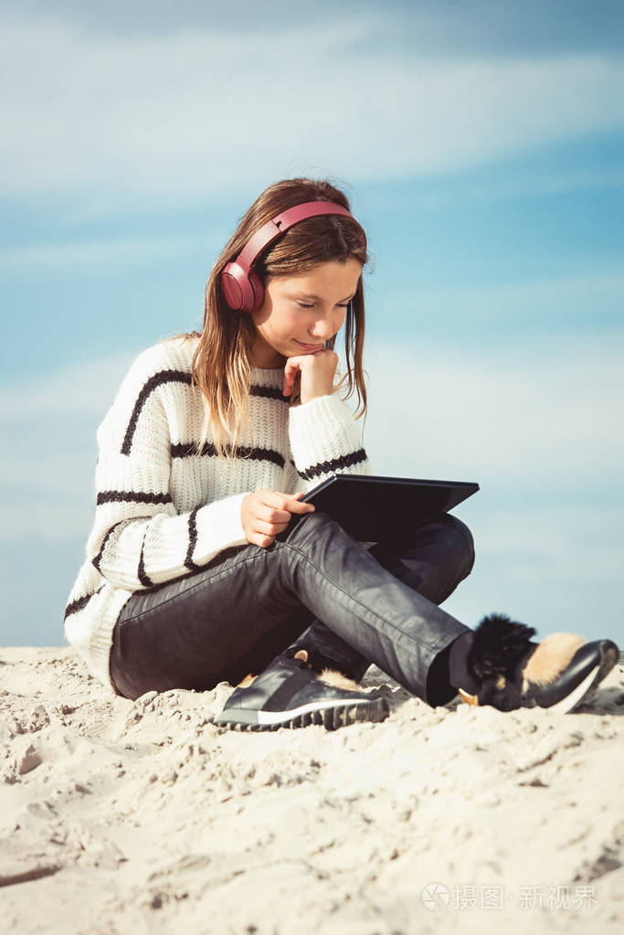 1 1岁的女孩戴着无线耳机, 在海滩上听着智能手机上的音乐.对着蓝天.