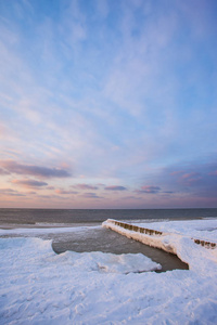 日落时分，在寒冷的冰雪覆盖的海面附近的冬季海滩上，积雪覆盖的防波堤