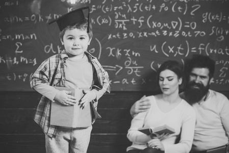 父母听他们的儿子, 检查错误与书黑板在背景。男孩向爸爸妈妈展示他的知识。聪明的孩子在毕业帽喜欢表演。准备考试概念
