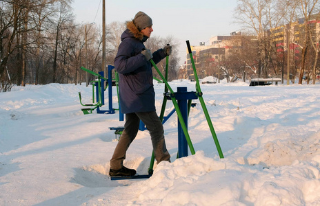 在一个冬季城市公园里, 身穿蓝色羽绒服的男子正在从事滑雪模拟器的工作。侧视图