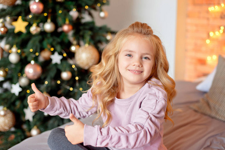 小美丽的卷发金发女孩坐在床上，在圣诞树的背景上微笑。 背景