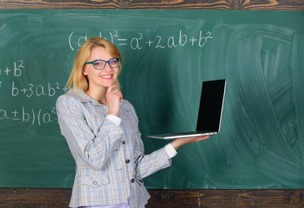 老师优雅的女士与现代笔记本电脑上网黑板背景。女教师戴眼镜拿着笔记本电脑上网。基础教育。远程教育理念