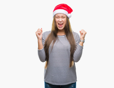 年轻美丽的高加索女人戴着圣诞帽，在孤立的背景下疯狂疯狂地大喊大叫，咄咄逼人的表情和手臂抬起。 挫折概念。