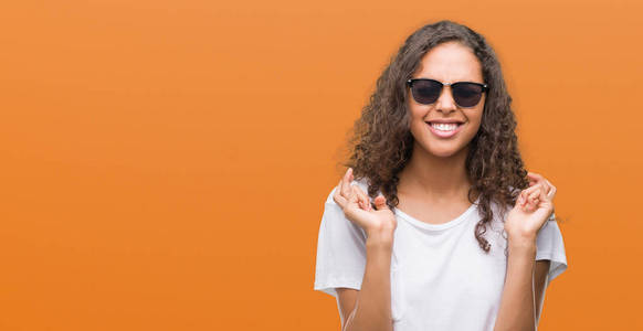 年轻的西班牙裔女人戴着太阳镜，微笑着交叉手指，充满希望，眼睛紧闭。 运气和迷信的概念。