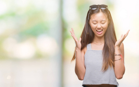 年轻的亚洲女人戴着太阳镜在孤立的背景上庆祝疯狂和疯狂的成功，举起手臂和闭上眼睛尖叫兴奋。 赢家概念