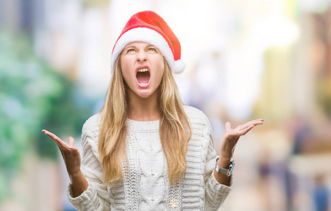 年轻漂亮的金发女人戴着圣诞帽，在孤立的背景上疯狂地大喊大叫，表情咄咄逼人，举起手臂。挫折的概念..