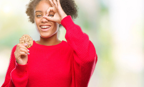 年轻的非洲裔美国妇女吃巧克力饼干，在孤立的背景下，快乐的脸微笑，做好的标志，手在眼睛看手指。
