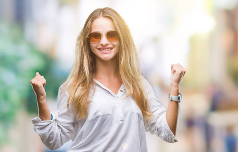 年轻漂亮的金发女人戴着太阳镜在孤立的背景上庆祝惊讶和惊讶的成功，举起手臂和睁开眼睛。赢家概念。