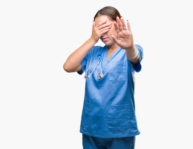 年轻的白种人医生女士穿着医疗制服，在孤立的背景上用手遮住眼睛，用悲伤和恐惧的表情做停止手势。尴尬和消极的概念。