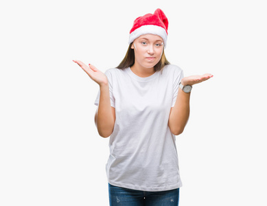 年轻美丽的白种人女人戴着圣诞帽，在孤立的背景上毫无头绪，表情混乱，举起手臂和手。怀疑的概念。