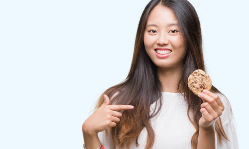 年轻的亚洲女人在孤立的背景上吃巧克力饼干，惊讶的脸指着自己