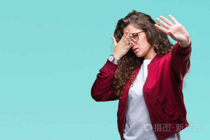 美丽的黑发卷发年轻女孩穿着夹克和眼镜在孤立的背景上用手遮住眼睛用