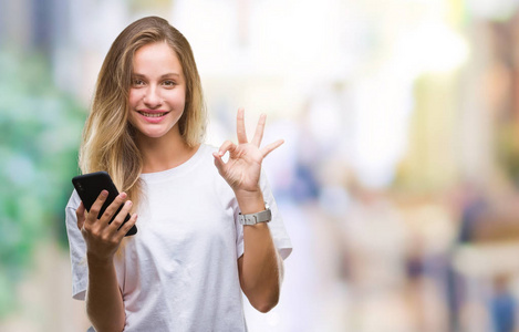 年轻漂亮的金发女人使用智能手机在孤立的背景下做OK标志手指优秀的符号