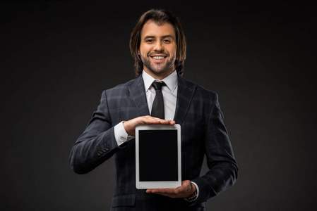 年轻英俊的商人拿着空白屏幕的数码平板电脑，微笑着看着与黑色隔离的相机