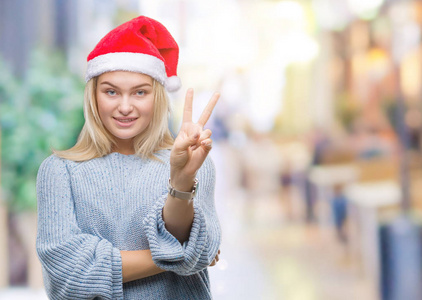 年轻的高加索女人戴着圣诞帽，在孤立的背景上微笑，快乐的脸对着镜头眨着眼睛，做着胜利的标志。 二号。