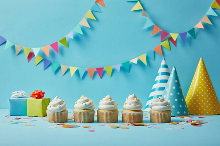 美味的纸杯蛋糕，糖洒，派对帽和礼物，蓝色背景，彩色发髻。