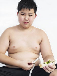 一个胖男孩一边吃三明治，一边检查他的腰围不满意