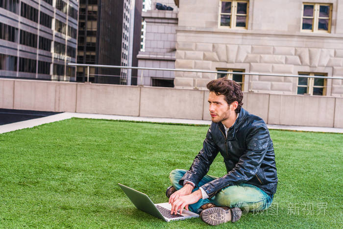 在纽约学习的欧洲研究生。穿着黑色皮夹克，蓝色牛仔裤，靴鞋，有胡子的年轻人，交叉腿，坐在绿色的草坪上，在笔记本电脑上工作，思考