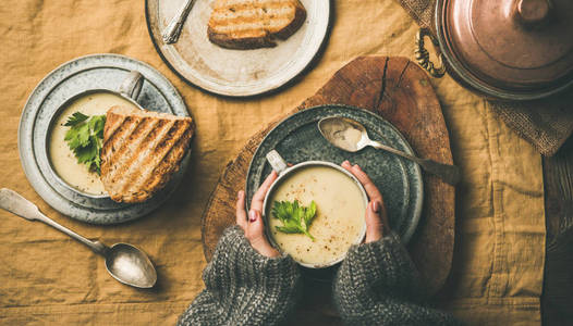 秋天的冬天回家晚餐。 平躺的秋季暖芹菜奶油汤在杯子，烤面包和女性的手在亚麻桌布的顶部视图。 舒适食物素食健康慢食概念
