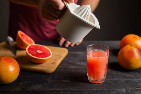 新鲜挤压的柚子汁是从手动榨汁机倒入玻璃杯中。 男性手中的白色塑料榨汁机。 在黑色的木桌上是完整的和切片的葡萄果实。