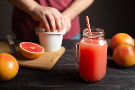 新榨的葡萄柚汁放在一个有手柄的罐子里。 雄性手在白色榨汁机上挤压水果。 在黑色的木桌上是完整的和切片的葡萄果实。