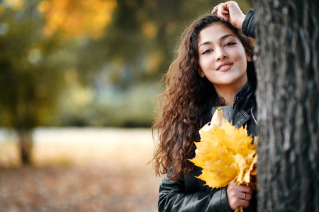 秋天城市公园里，有黄叶的女人站在大树附近。 肖像特写。