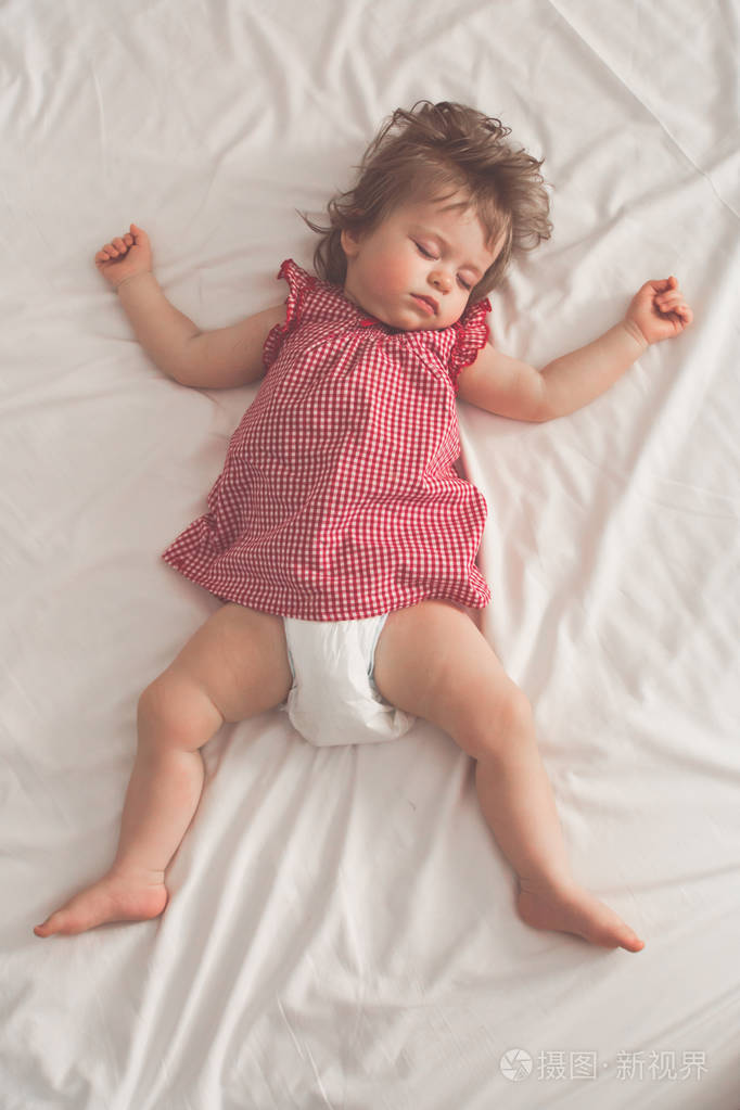 小女孩仰卧,张开双臂,没有奶嘴,躺在一张白色床单的床上.
