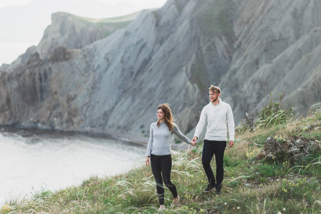 年轻夫妇走在北欧海岸，在春天的山景中，穿着休闲风格的衣服毛衣和牛仔裤。