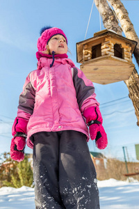 可爱的白种人女孩穿着运动冬季夹克，在户外玩雪。 背景上树上的鸟类喂食器。 寒假和假期的概念。 儿童户外活动。