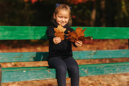小女孩坐在长凳上看树叶。她收集树叶, 带着。秋天森林