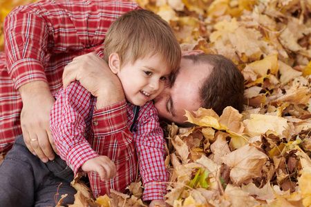 父子俩躺在黄叶上，在秋天的城市公园里玩得很开心。他们摆姿势，微笑，玩耍。明亮的黄树。