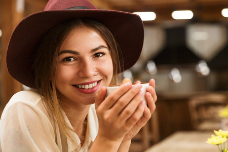 一个戴着帽子的快乐女孩坐在室内的咖啡桌旁，端着一杯茶