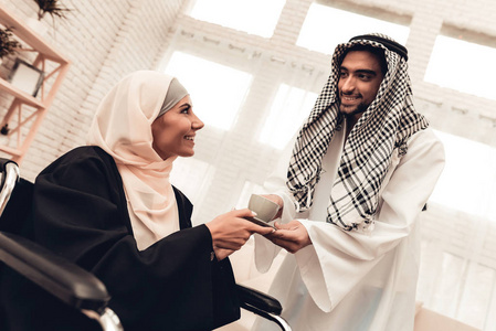 年轻的阿拉伯男人在轮椅上给妻子送杯子。 阿拉伯家庭的概念。 坐在轮椅上。 残疾妇女。 喝茶的女人。 坐着的穆斯林女人。 一杯咖啡
