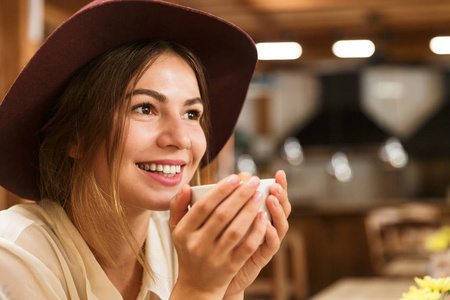 一个戴着帽子的漂亮女孩坐在咖啡桌旁，手里拿着一杯茶
