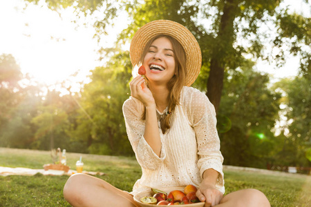 微笑着戴着夏帽的年轻女孩坐在草地上野餐，吃着碗里的水果