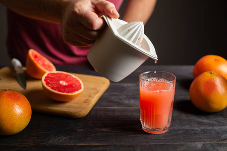 新鲜挤压的柚子汁是从手动榨汁机倒入玻璃杯中。 男性手中的白色塑料榨汁机。 在黑色的木桌上是完整的和切片的葡萄果实。