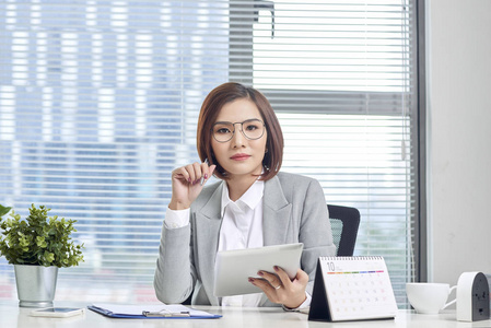 自信的亚洲商界女性坐在办公桌前使用数码平板电脑的画像