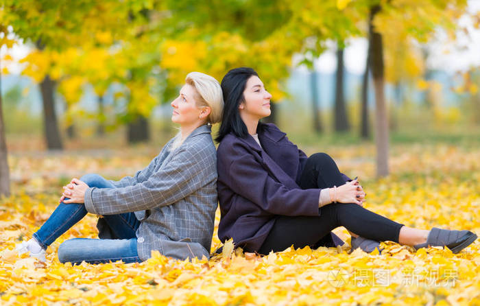 两个年轻的女人背靠背坐在公园里,地上长满了明亮的黄色叶子,从侧面看