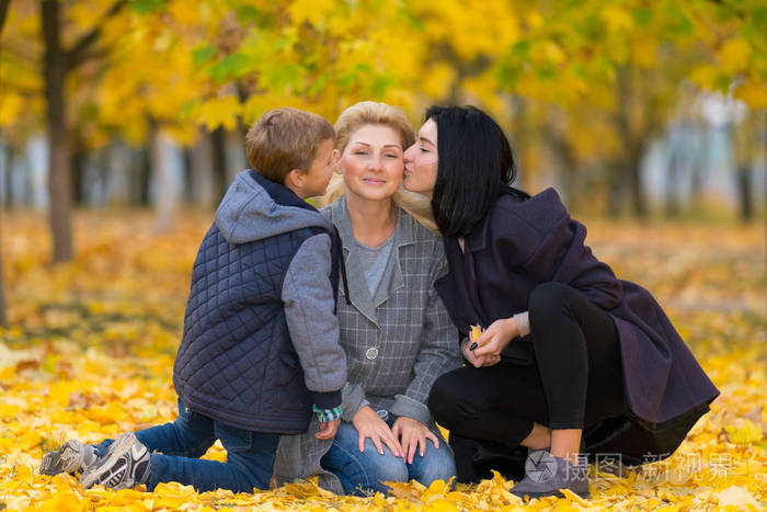 小男孩和他的姐姐在明亮的秋日坐在公园的地上在明亮的黄叶上亲吻妈妈